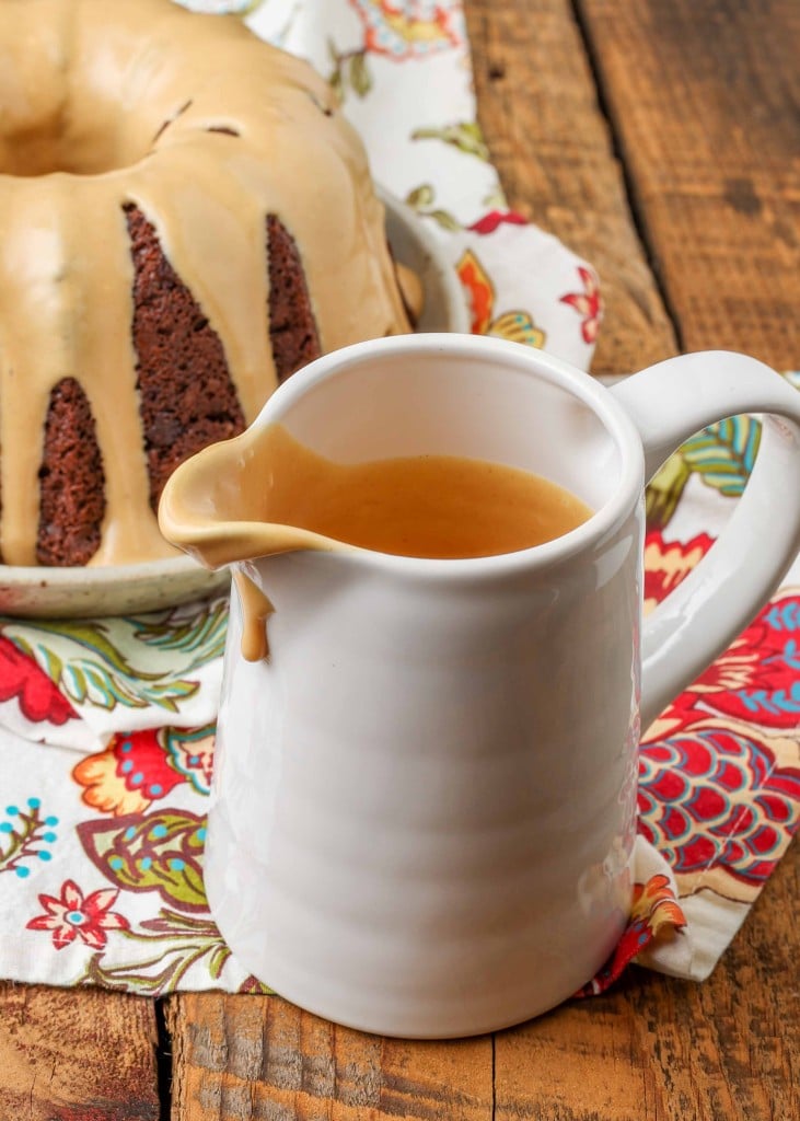 a used white pitcher stands on top of a colorful tea towel next to the bundt cake it has just finished glazing.