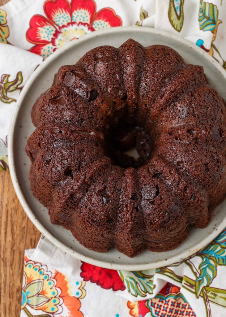 A top down shot of a chocolate bundt cake on a speckled white plate with a colorful tea towel peeking out from beneath the plate