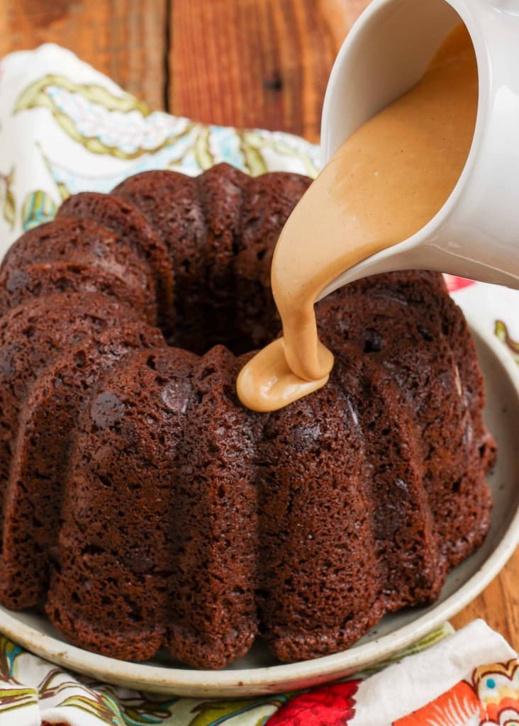luscious peanut butter glaze pours thickly out of a white pitcher onto the top of a chocolate bundt cake on a speckled white plate
