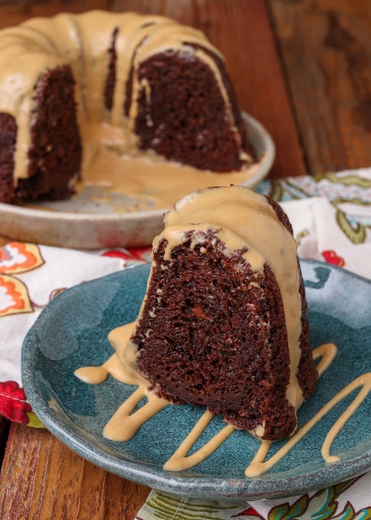 A slice of chocolate bundt cake drizzled with peanut butter glaze on a blue ceramic plate with the rest of the cake visible in the background.