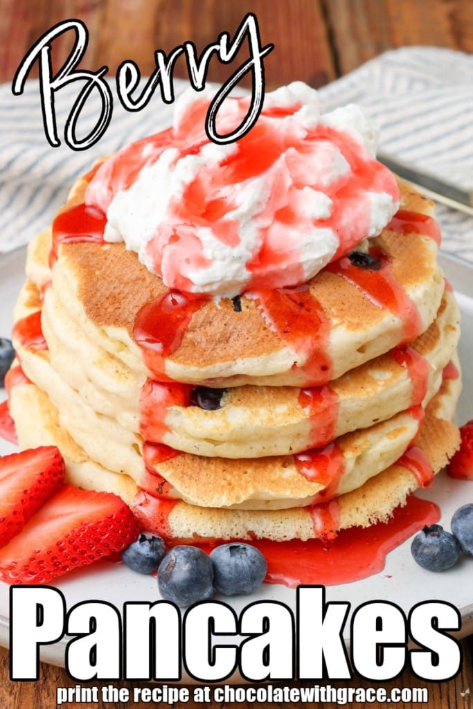 pancakes made with blueberries and strawberries on a plate, surrounded by more berries, topped with whipped cream and strawberry syrup