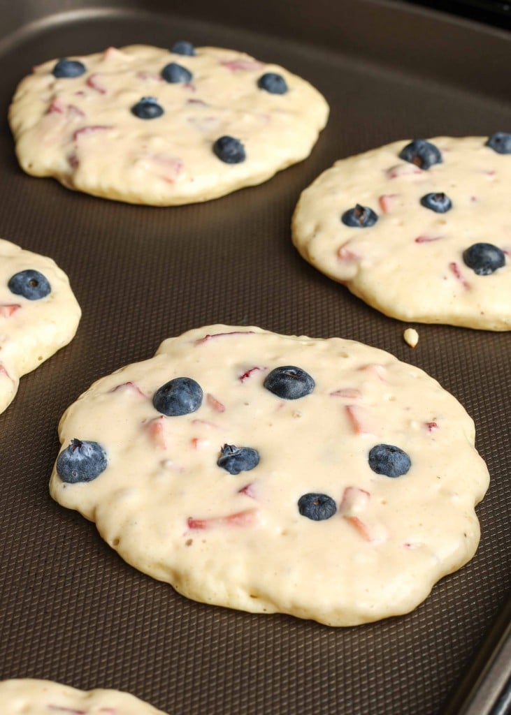 pancakes cooking on a griddle, with fresh blueberries sitting on top of the batter, and chunks of strawberry visible within