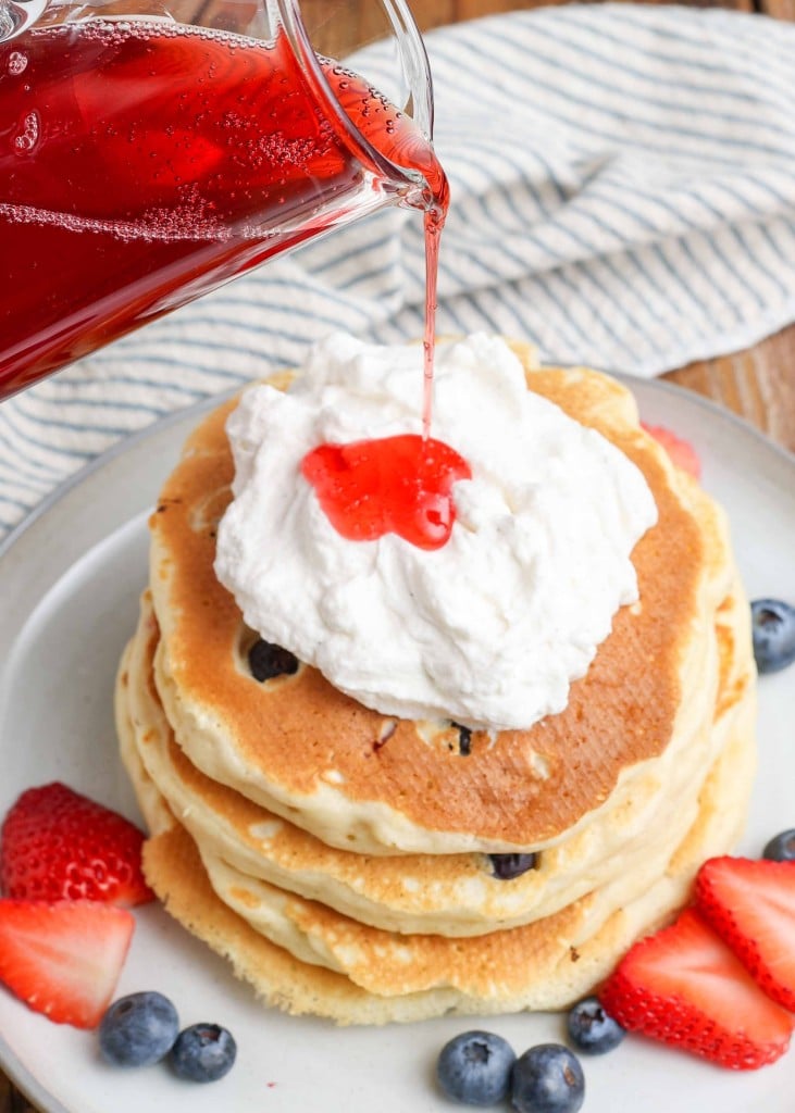 a clear pitcher pours strawberry syrup onto a dollop of whipped cream atop a stack of pancakes with strawberry and blueberries