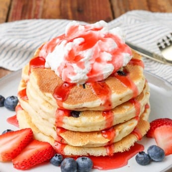 a plate full of mixed berry pancakes, ready to eat on a white plate, with syrup and whipped cream
