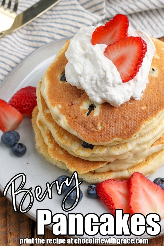 a stack of fluffy pancakes made with blueberries and strawberries on a plate, surrounded by more berries, topped with whipped cream and strawberry syrup
