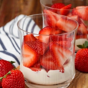 strawberries rest atop a layer of fluffy sweet cream in the bottom of a ball glass with more strawberries visible in the background