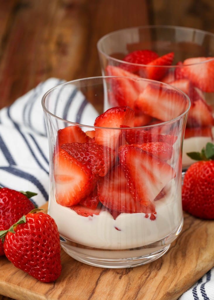 strawberries rest atop a layer of fluffy sweet cream in the bottom of a ball glass with more strawberries visible in the background
