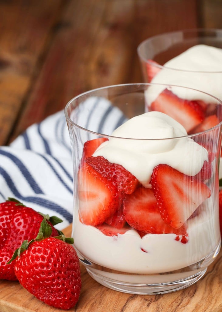 A short glass is filled partway with smooth, fluffy whipped cream and halved strawberries with a blue striped towel in the background on a wooden tabletop