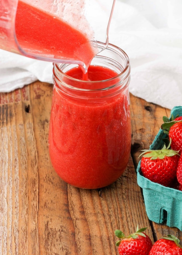 Strawberry puree pouring into a glass jar with basket of fresh berries