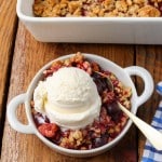 Dark red cherry oatmeal crisp topped with white vanilla ice cream in a white bowl with a silver spoon and checkered blue towel