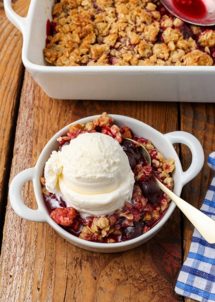 Dark red cherry oatmeal crisp topped with white vanilla ice cream in a white bowl with a silver spoon and checkered blue towel