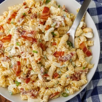 Overhead vertical shot of bacon pasta salad, served in a white bowl with a checkered blue and white towel and a silver fork
