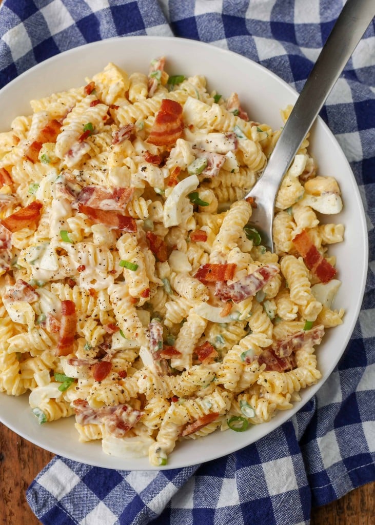 Overhead vertical shot of bacon pasta salad, served in a white bowl with a checkered blue and white towel and a silver fork