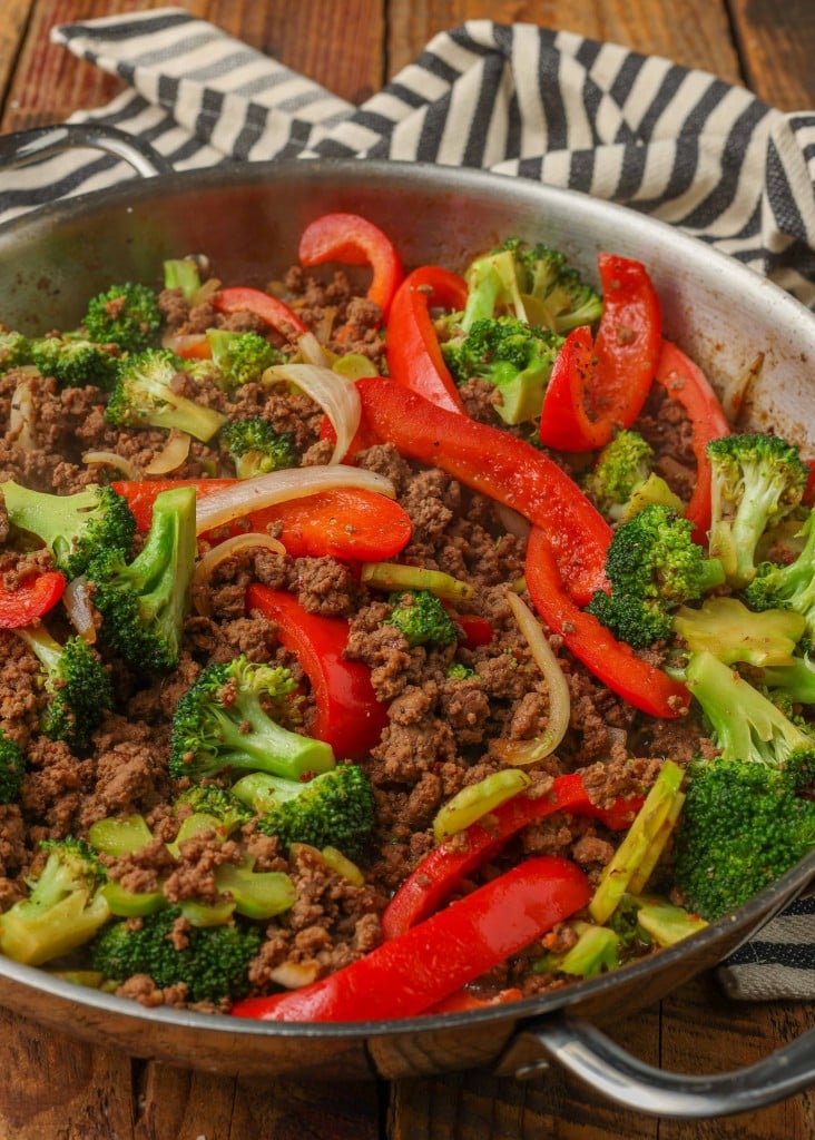Overhead horizontal shot of ground beef, broccoli, and bell pepper stir fry in a stainless steel skillet, next to a striped gray and white towel