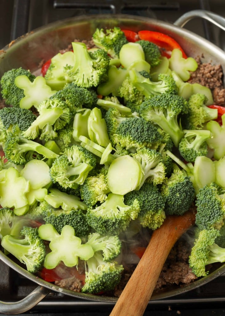 Overhead shot of ground beef, red bell peppers, yellow onions, and broccoli cooking in a stainless steel skillet