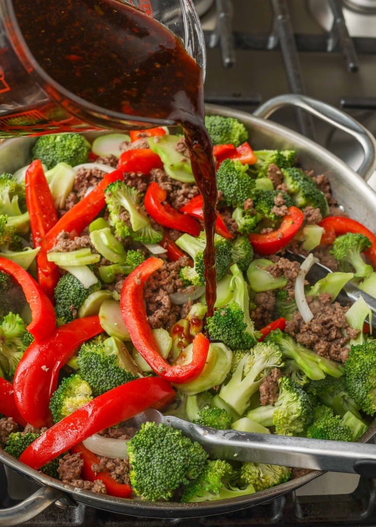 Overhead shot of sauce being poured into ground beef, red bell peppers, yellow onions, and broccoli cooking in a stainless steel skillet
