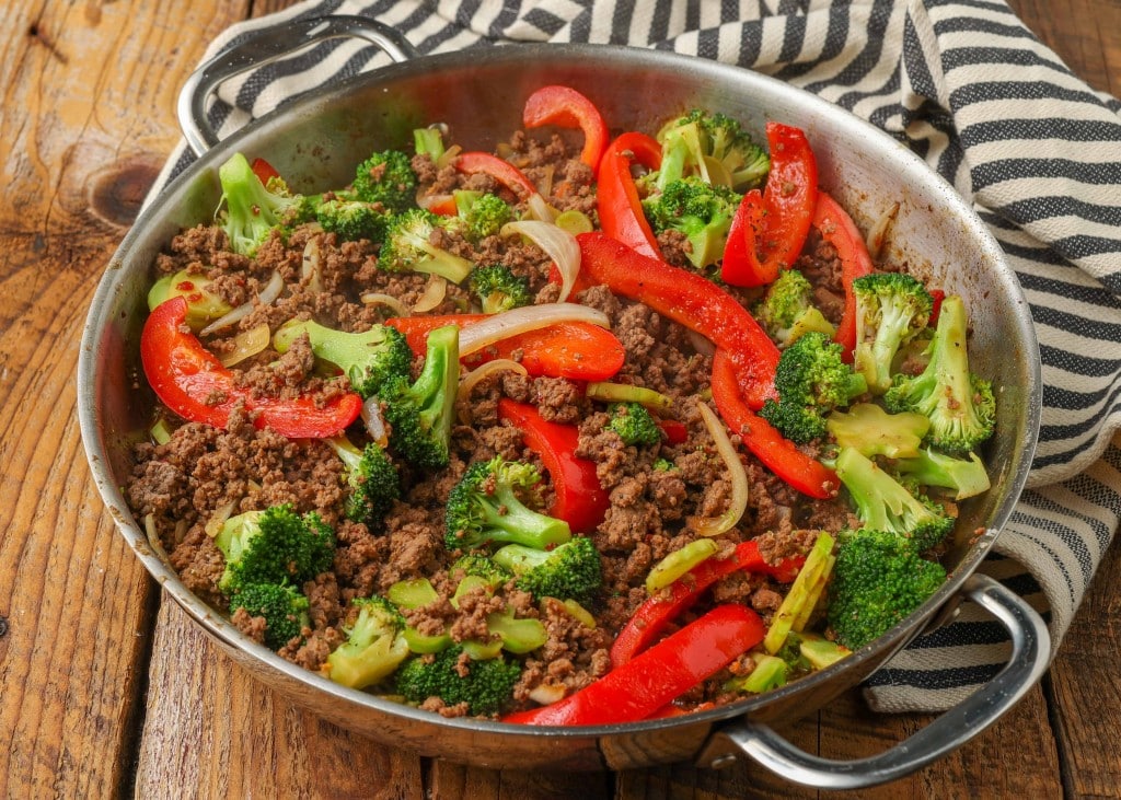 Overhead horizontal shot of ground beef, broccoli, and bell pepper stir fry in a stainless steel skillet, next to a striped gray and white towel