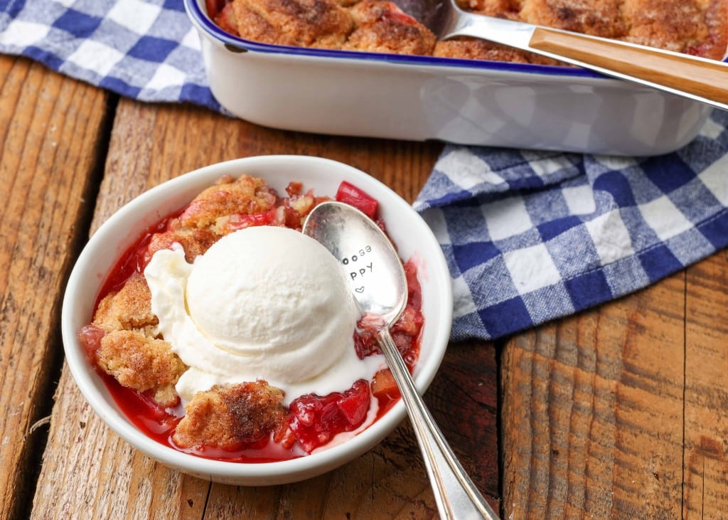 Overhead shot of golden rhubarb cobbler fresh from the pan, topped with vanilla ice cream, served in a white bowl with a checkered blue and white hand towel and a silver spoon