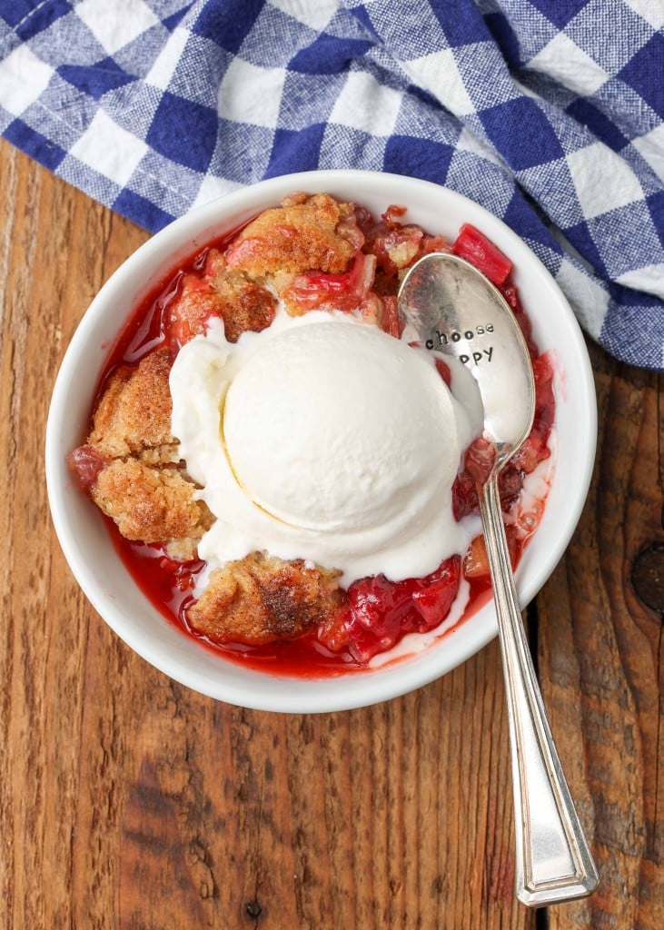 Overhead shot of a silver spoon digging into the golden crust of rhubarb cobbler fresh from the oven, topped with a scoop of vanilla ice cream, served in a white bowl on a checkered blue and white hand towel