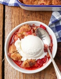 Overhead shot of a silver spoon digging into the golden crust of rhubarb cobbler fresh from the oven, topped with a scoop of vanilla ice cream, served in a white bowl on a checkered blue and white hand towel