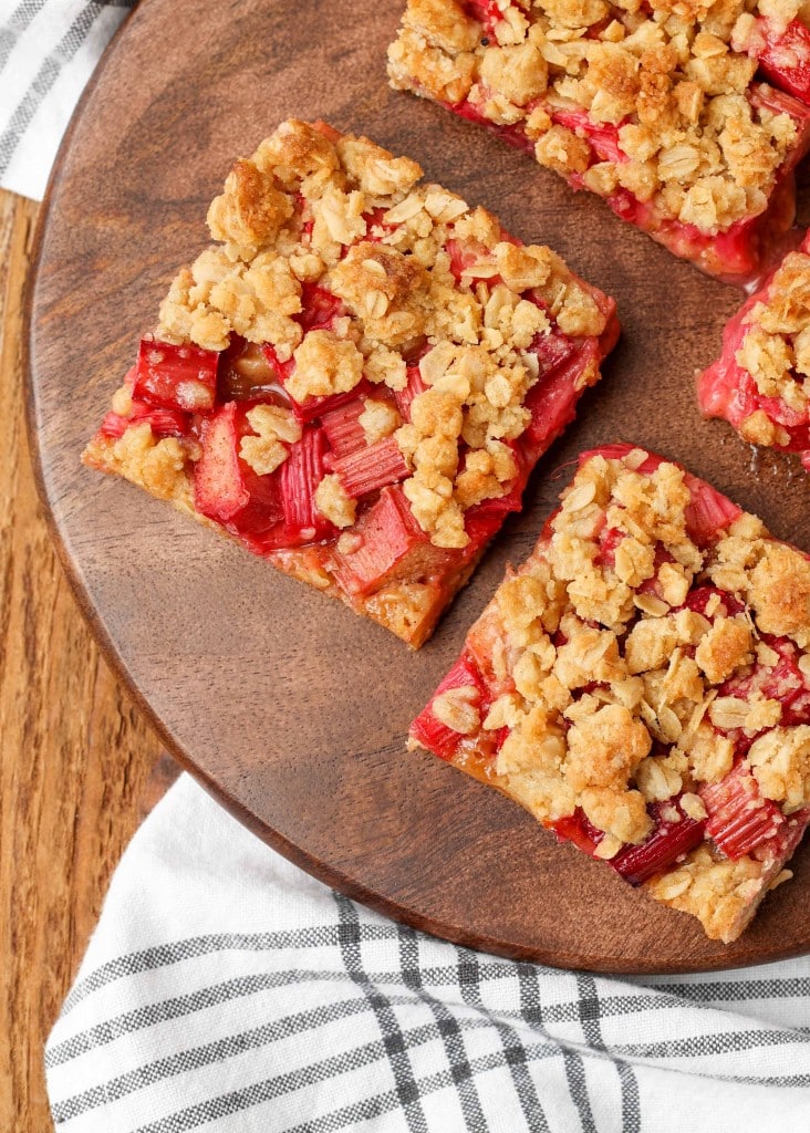 Overhead shot of rhubarb bars with crunchy oatmeal topping, served on a round wooden cutting board with a striped gray and white towel