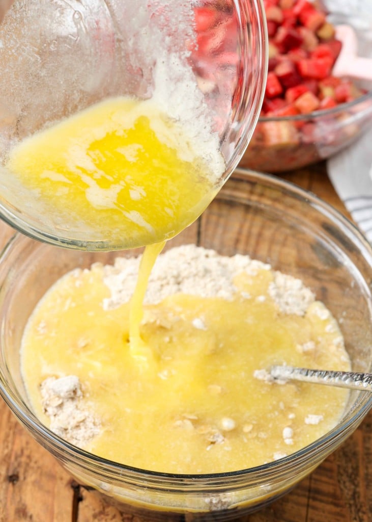 Close-up shot of melted butter being poured into a glass bowl filled with chopped rhubarb and dried ingredients