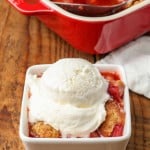 a scoop of white vanilla ice cream has been placed atop a serving of strawberry rhubarb cobbler in a white dish on a wooden tabletop