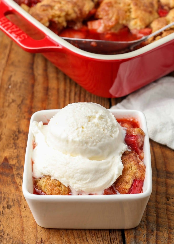 a scoop of white vanilla ice cream has been placed atop a serving of strawberry rhubarb cobbler in a white dish on a wooden tabletop