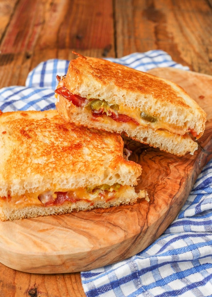artfully arranged halves of a bacon grilled cheese on top of a wooden serving board with a blue and white checkered napkin visible in the background