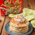 Fruity pebbles pancakes stacked on a blue plate, topped with whipped cream and a sprinkling of fruity pebbles cereal