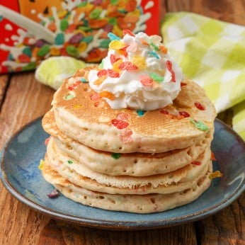 Fruity pebbles pancakes stacked on a blue plate, topped with whipped cream and a sprinkling of fruity pebbles cereal