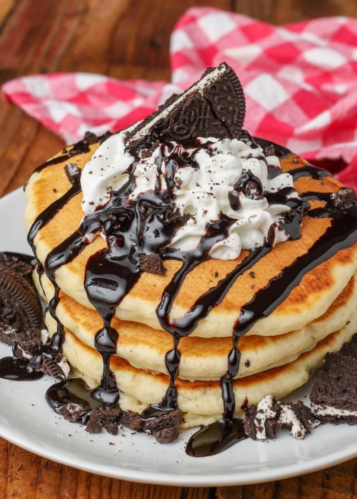 A stack of three oreo pancakes on a white plate with whipped cream and chocolate syrup