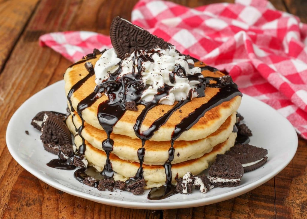 a stack of fluffy oreo pancakes, topped with whipped cream, drizzled with chocolate sauce, and surrounded by oreo pieces on the rim of the white plate.