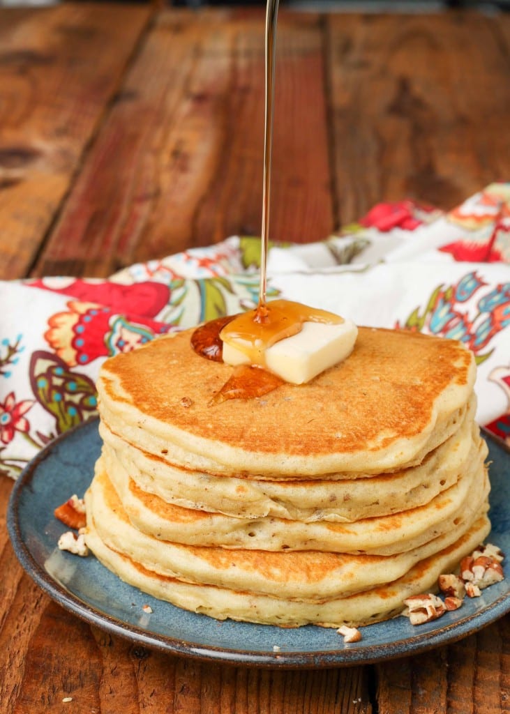 a pour shot of syrup being drizzled over a pat of butter atop a stack of pancakes