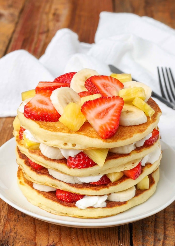 A vertically aligned photo of pancakes that has been layered with whipped cream, bananas, strawberries, and pineapple tidbits.