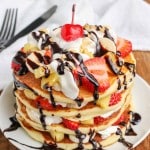 A vertically aligned photo of a large stack of pancakes on a white plate, topped with sliced bananas, strawberries, pineapple bits, whipped cream, and a drizzle of chocolate sauce.