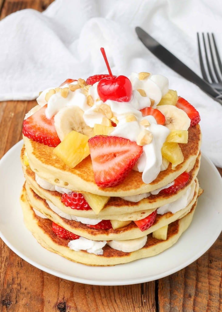 A vertically aligned photo of pancakes that has been layered with whipped cream, bananas, strawberries, and pineapple tidbits.