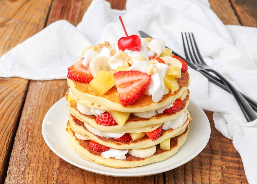 A horizontally aligned photo of pancakes that has been layered with whipped cream, bananas, strawberries, and pineapple tidbits.