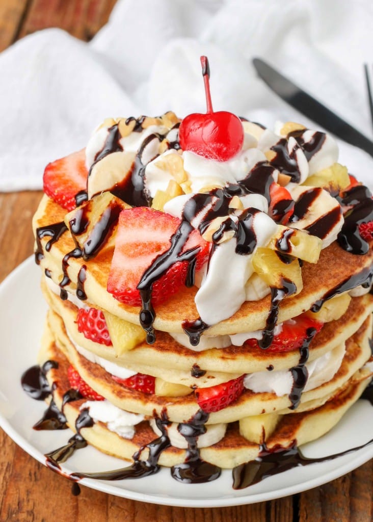 A cherry has been placed atop this stack of banana split pancakes with whipped cream and chocolate sauce.