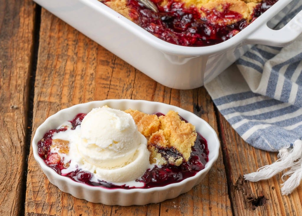 a horizontally aligned photo of an oblong dish with a serving of blueberry dump cake in it, topped with vanilla ice cream, on a wooden tabletop.