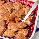 a metal spoon has been inserted into the blueberry peach cobbler, lifting out a serving of the warm dessert.
