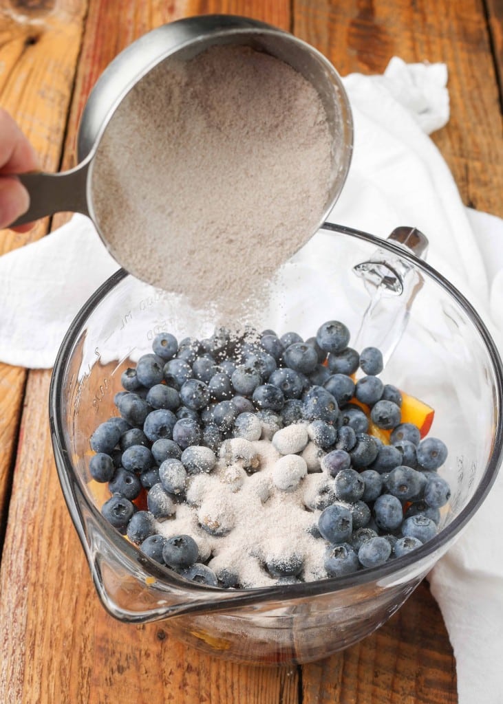 Sprinkling sugar over the blueberries and peaches in a glass mixing bowl