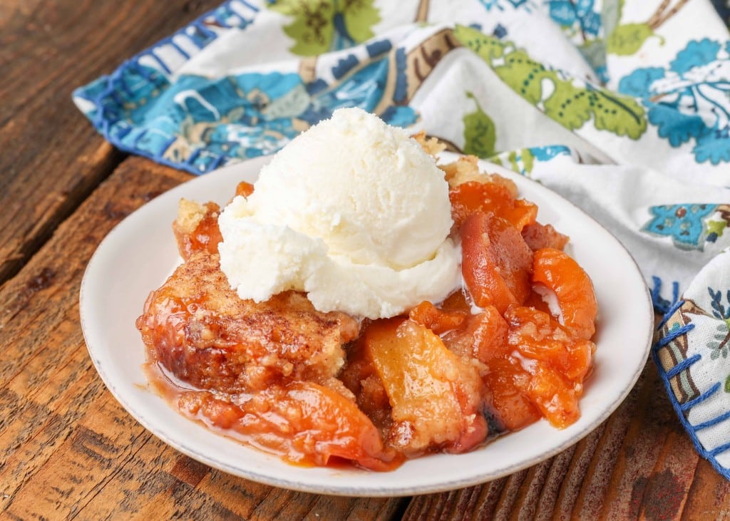 An individual serving of crockpot peach cobbler on a white plate on a wooden tabletop with a colorful blue towel behind it.