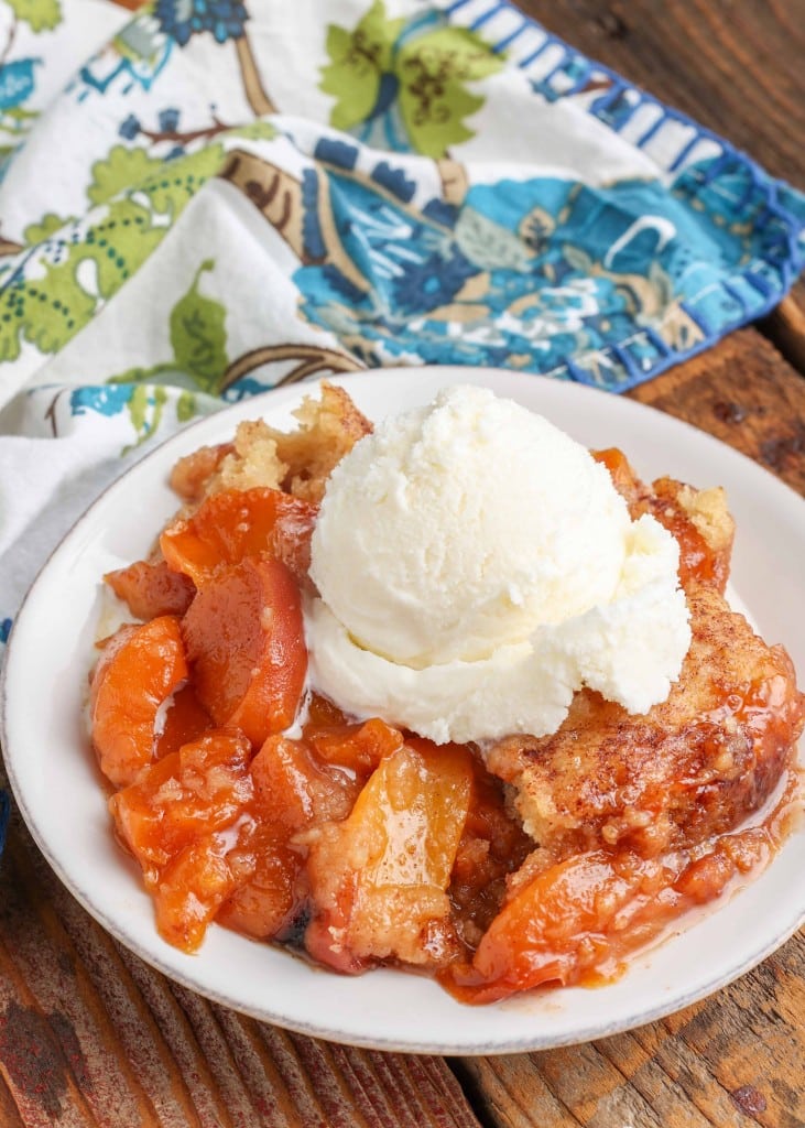 A serving of crockpot peach cobbler is visible on a small white plate with a scoop of ice cream.
