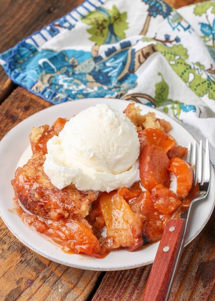 A fork with a wooden handle is visible on a white plate beside a serving of peach cobbler with a scoop of vanilla ice cream.