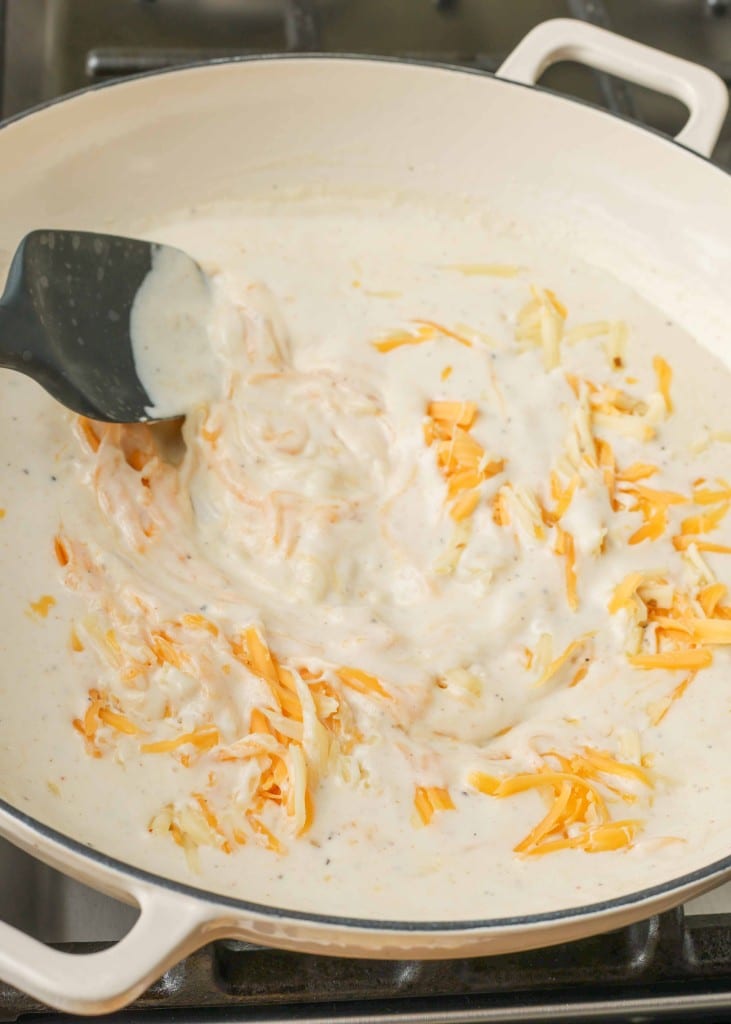 A black spatula is used to fold in the shredded cheese into the creamy sauce in the pan.