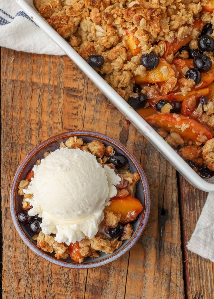 a vertically aligned photo of a serving of peach crisp beside the baking pan full of it. the smaller serving has been topped with ice cream.