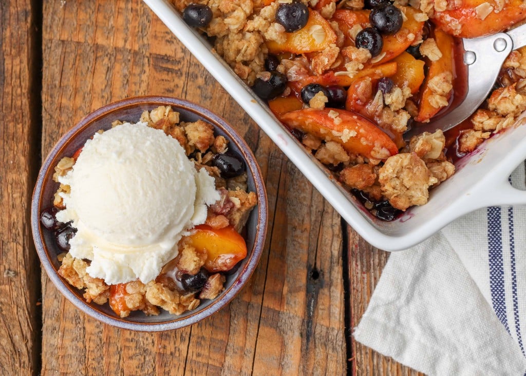 a horizontally aligned photo of a serving of peach crisp beside the baking pan full of it. the smaller serving has been topped with ice cream.
