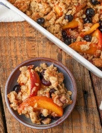 a serving of peach blueberry crisp in a light blue ceramic bowl on a wooden tabletop, beside the baking pan full of the rest of the crisp
