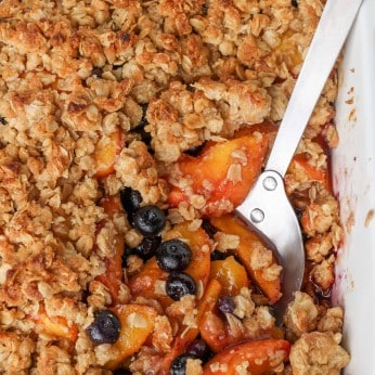 sliced peaches and blueberries are visible beneath the oat crust of the crisp in this top down shot of the pan with a metal spoon in it.